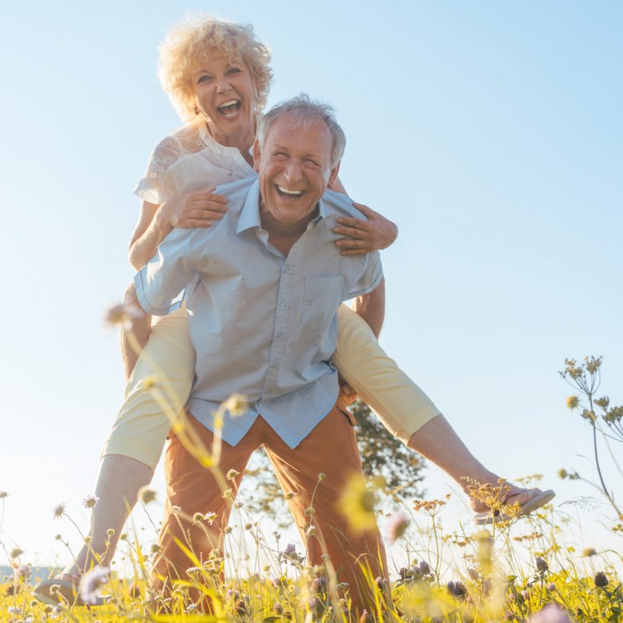 Low-angle view portrait of a happy senior man laughing while carrying his partner on his back in a sunny day of summer in the countryside