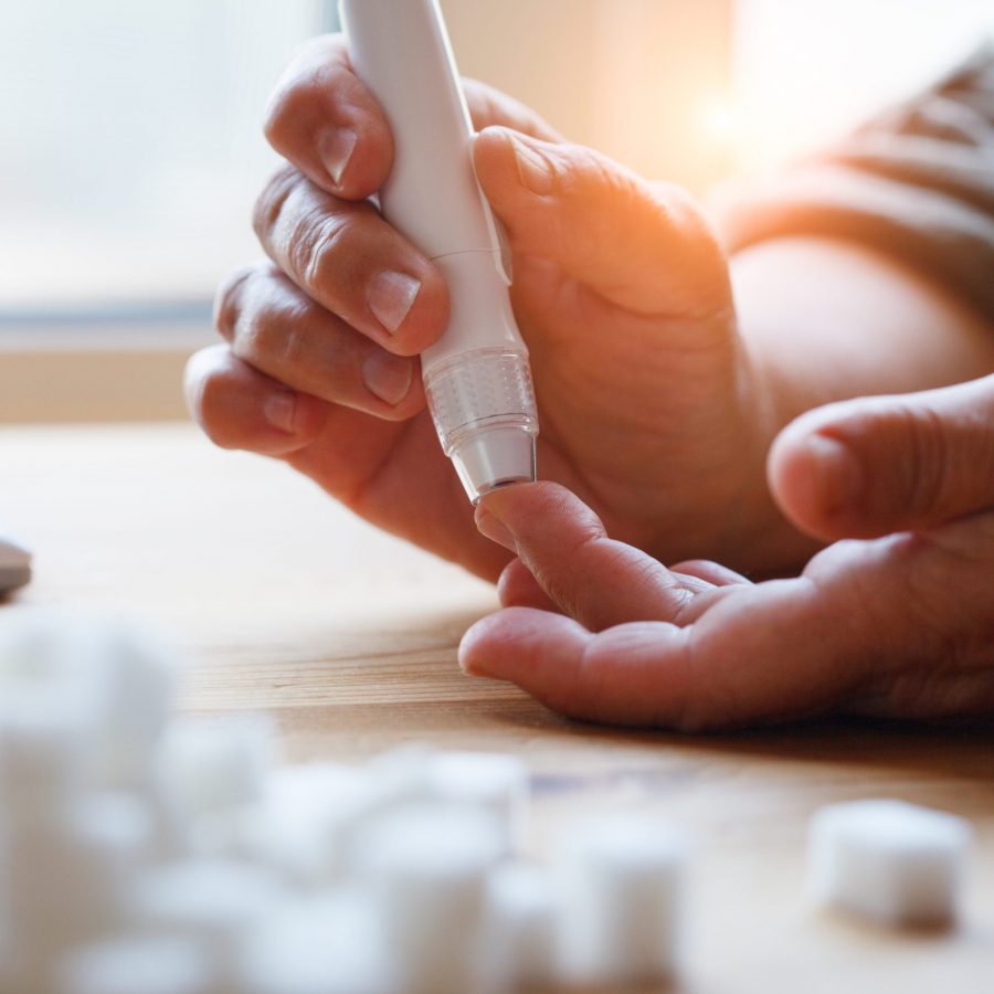 senior woman hands using lancet on finger at home to check blood sugar level, glucometer  and sugar cubes on wooden table close up, diabetes concept,  elderly health care, sunny morning